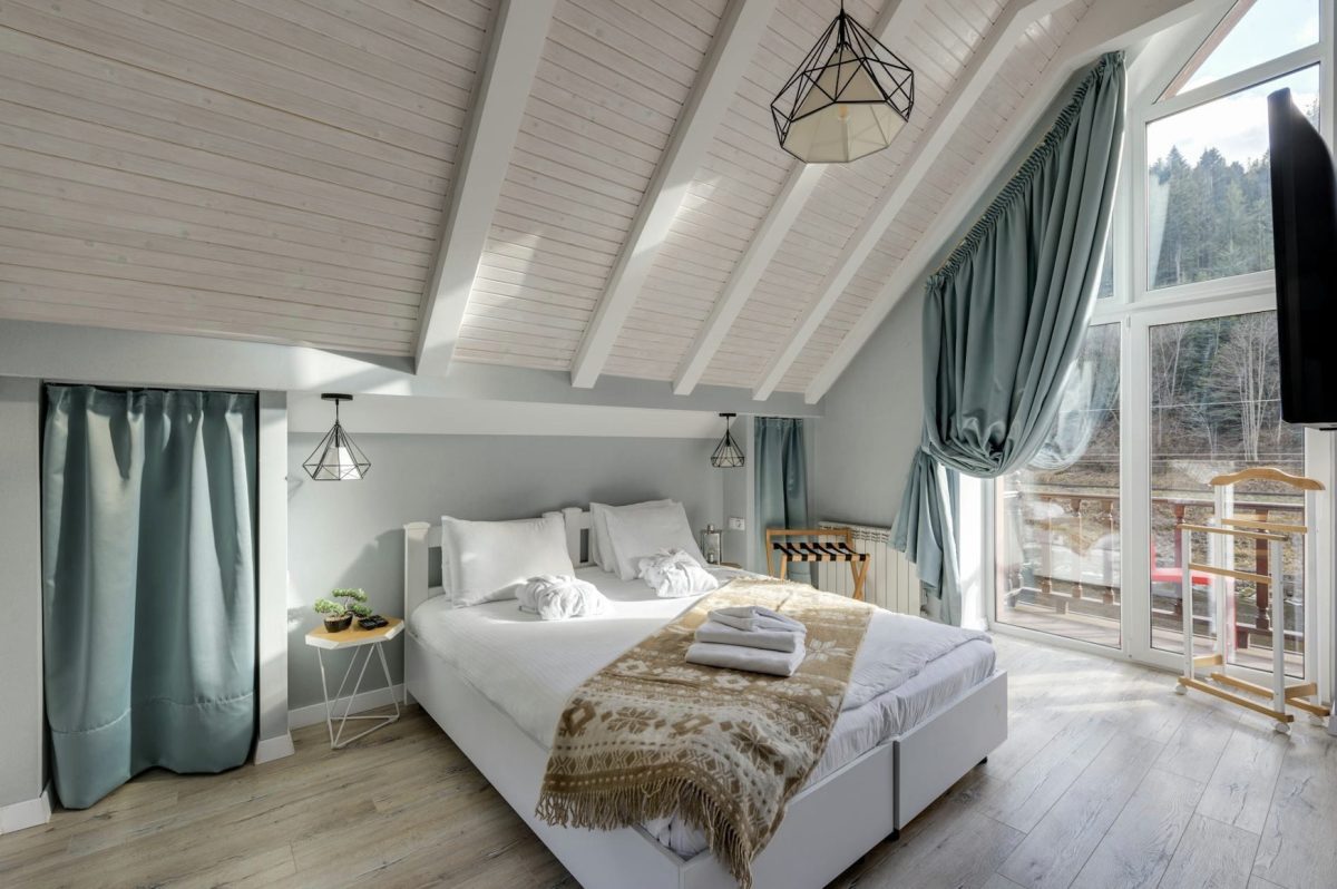 Cozy attic bedroom with a slanted wooden ceiling, large window, double bed, and folded towels. Drapes and a small plant add decorative touches.