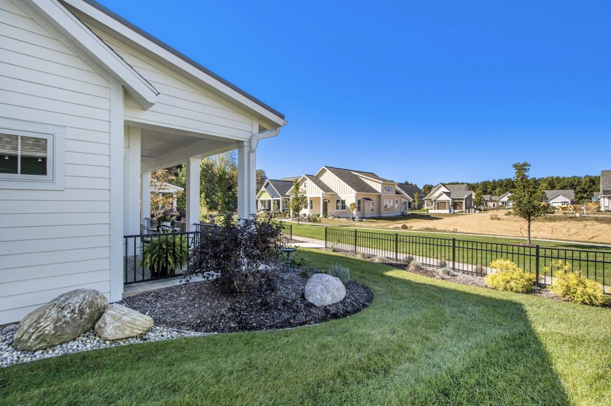 A suburban neighborhood with white houses, a manicured lawn, rocks, and a metal fence in the foreground under a clear blue sky.